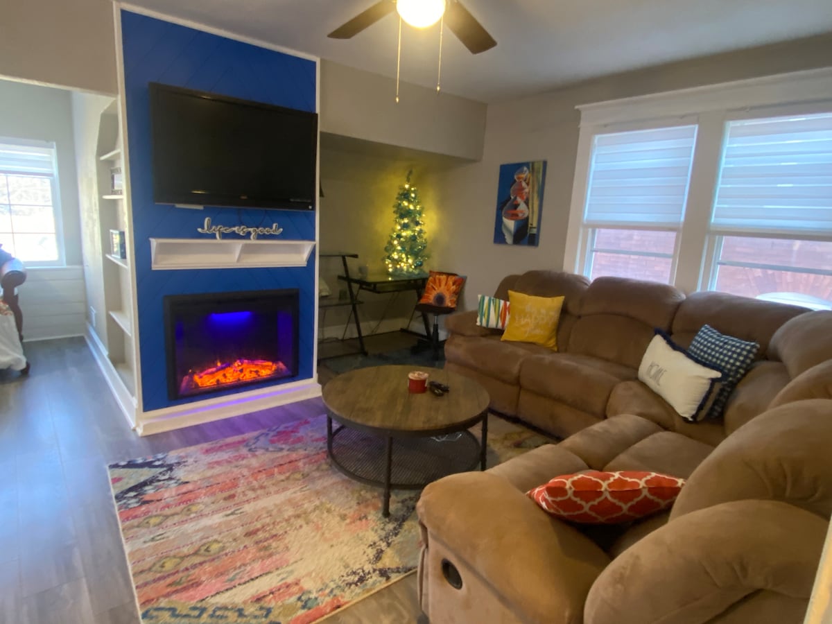A comfortable living area features a spacious sectional sofa with decorative pillows. A modern flat-screen TV is mounted above a glowing electric fireplace. Natural light is provided by two windows, and a small table sits nearby, accented by a festive miniature tree.