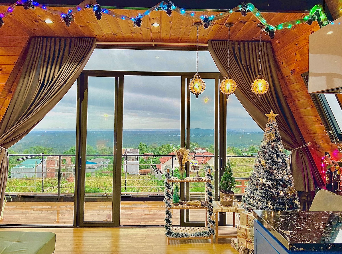 A modern living area features large glass doors that provide expansive views of the surrounding landscape. Decorative string lights are draped along the walls. A small, decorated Christmas tree and a stylish seating arrangement enhance the cozy ambiance of the space.