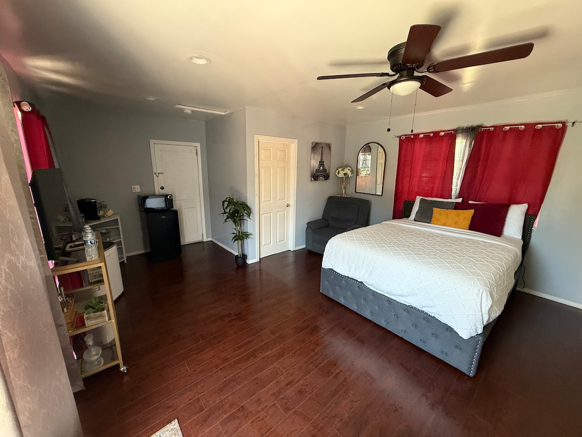 A private room features a queen-sized bed with crisp linens and a cozy twin bed alongside. A small work desk sits in the corner, and a flat-screen TV is mounted on the wall. Natural light enters through windows draped with red curtains, while a mini fridge and coffee machine are provided.
