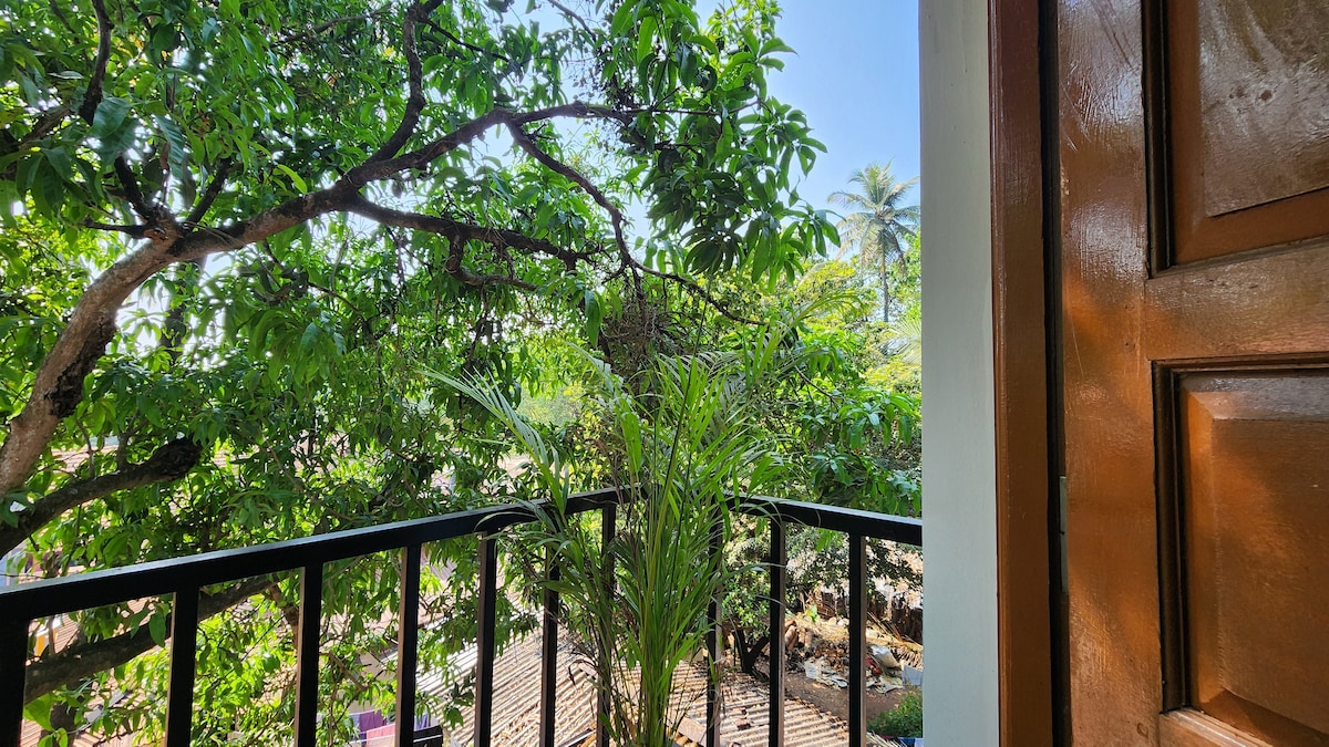 A private balcony is framed by lush greenery, including a tall palm and various trees, providing a natural view. A potted plant occupies the corner, while the sunlight filters through the leaves, creating a serene environment. The balcony railing is black, contrasting with the warm wooden door.