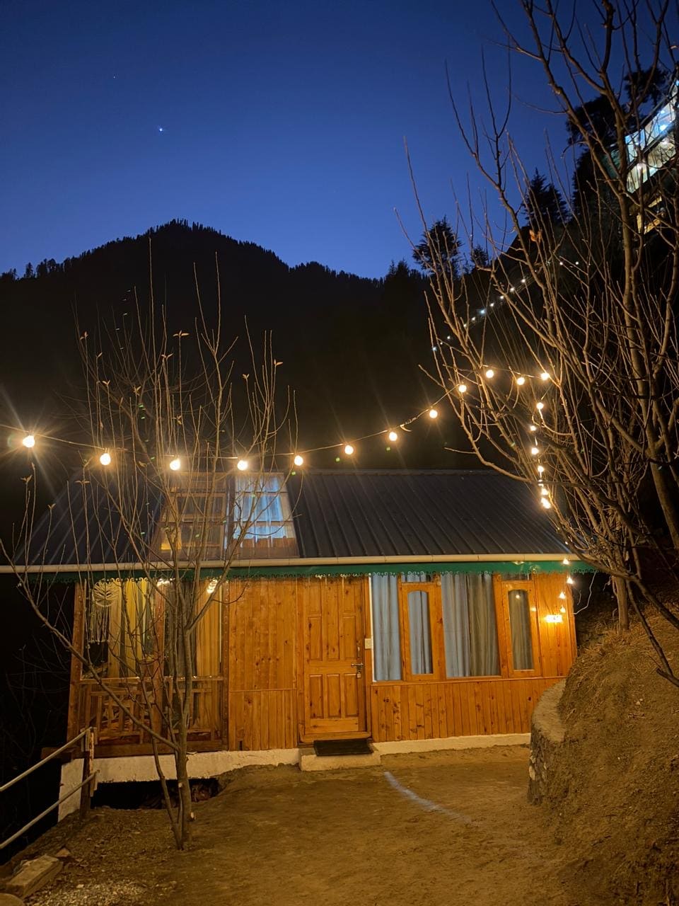 A charming wooden structure is illuminated by string lights, set against a night sky. Surrounding trees and a mountain backdrop create a serene atmosphere. The main entryway features glowing windows, reflecting the warmth inside, while the path leads towards the inviting entrance.