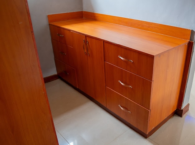 A wooden storage cabinet occupies a corner, featuring multiple drawers with sleek metallic handles. The warm tones of the wood contrast with the neutral walls and floor, offering an organized space for personal belongings.