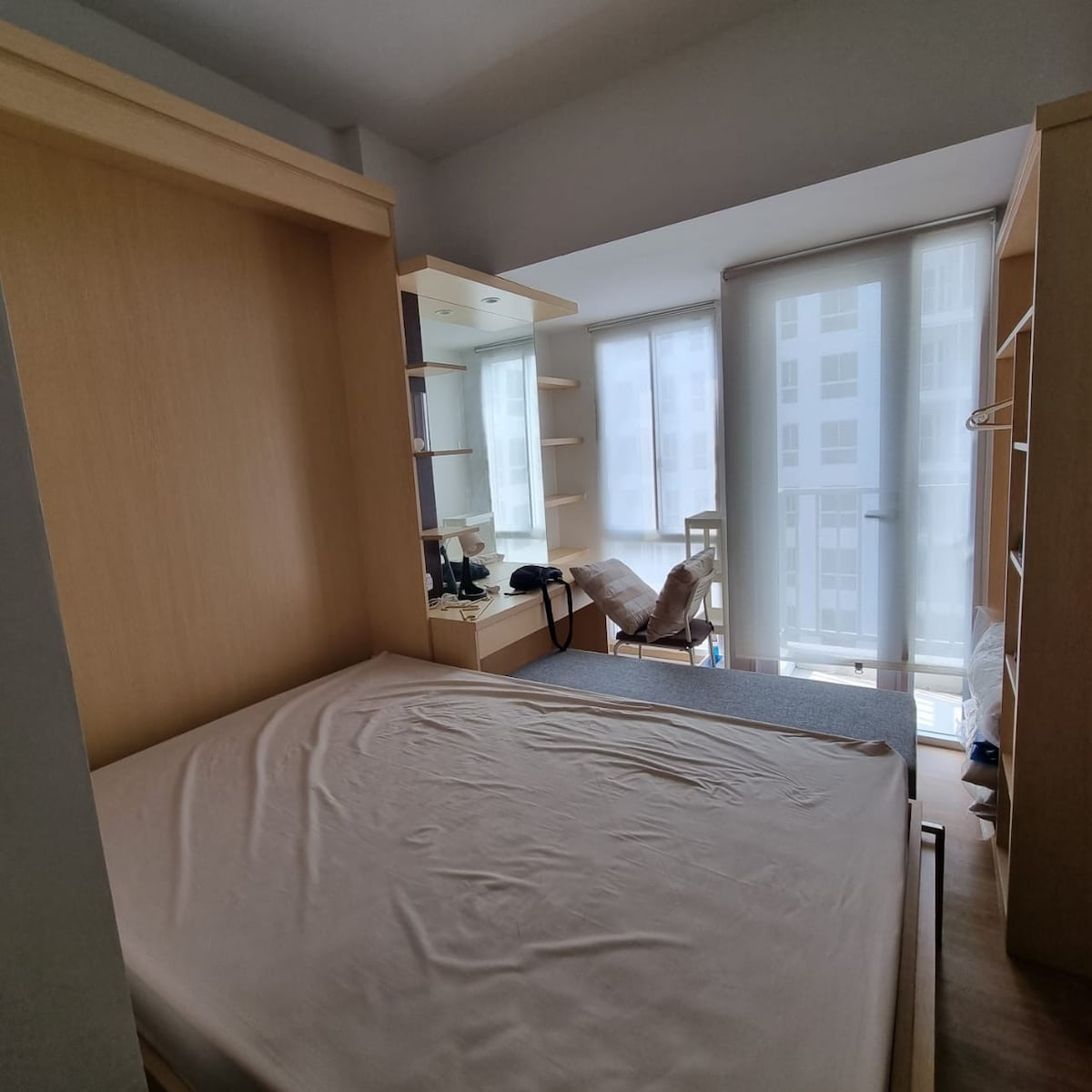 A cozy sleeping area features a folded-down bed with a plain mattress. Surrounding built-in shelves offer storage, while large windows allow natural light to fill the space. A chair is positioned nearby, and a table with lighting is also visible.
