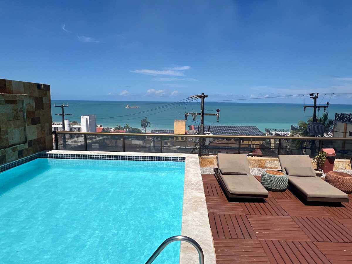 A rooftop area features a swimming pool with clear blue water, accompanied by two sun loungers positioned for relaxation. A panoramic view of the ocean and coastal landscape is visible in the background, framed by clear skies and distant horizons.
