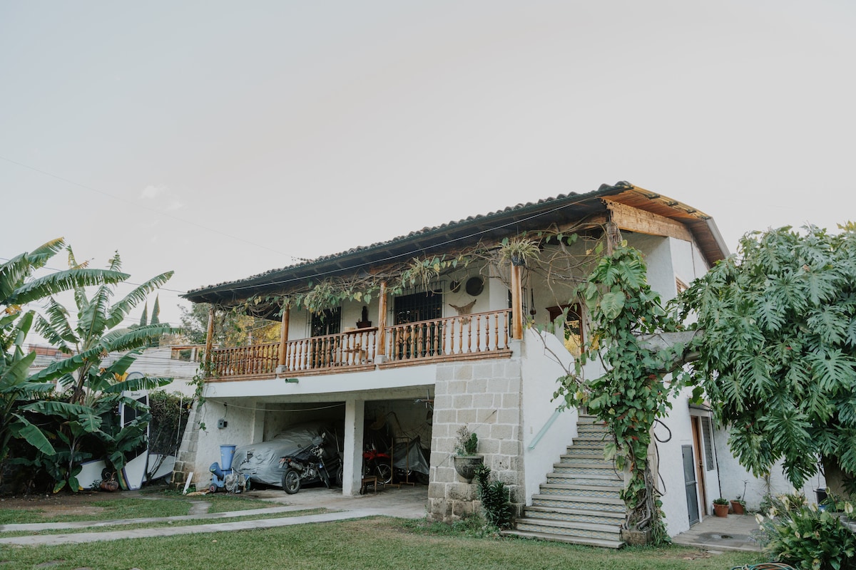 The exterior of a colonial-style building is presented, featuring a wraparound balcony adorned with wooden railings. Lush greenery surrounds the structure, while a staircase leads to the upper level. Vehicles are parked beneath the building, contributing to a sense of accessibility.