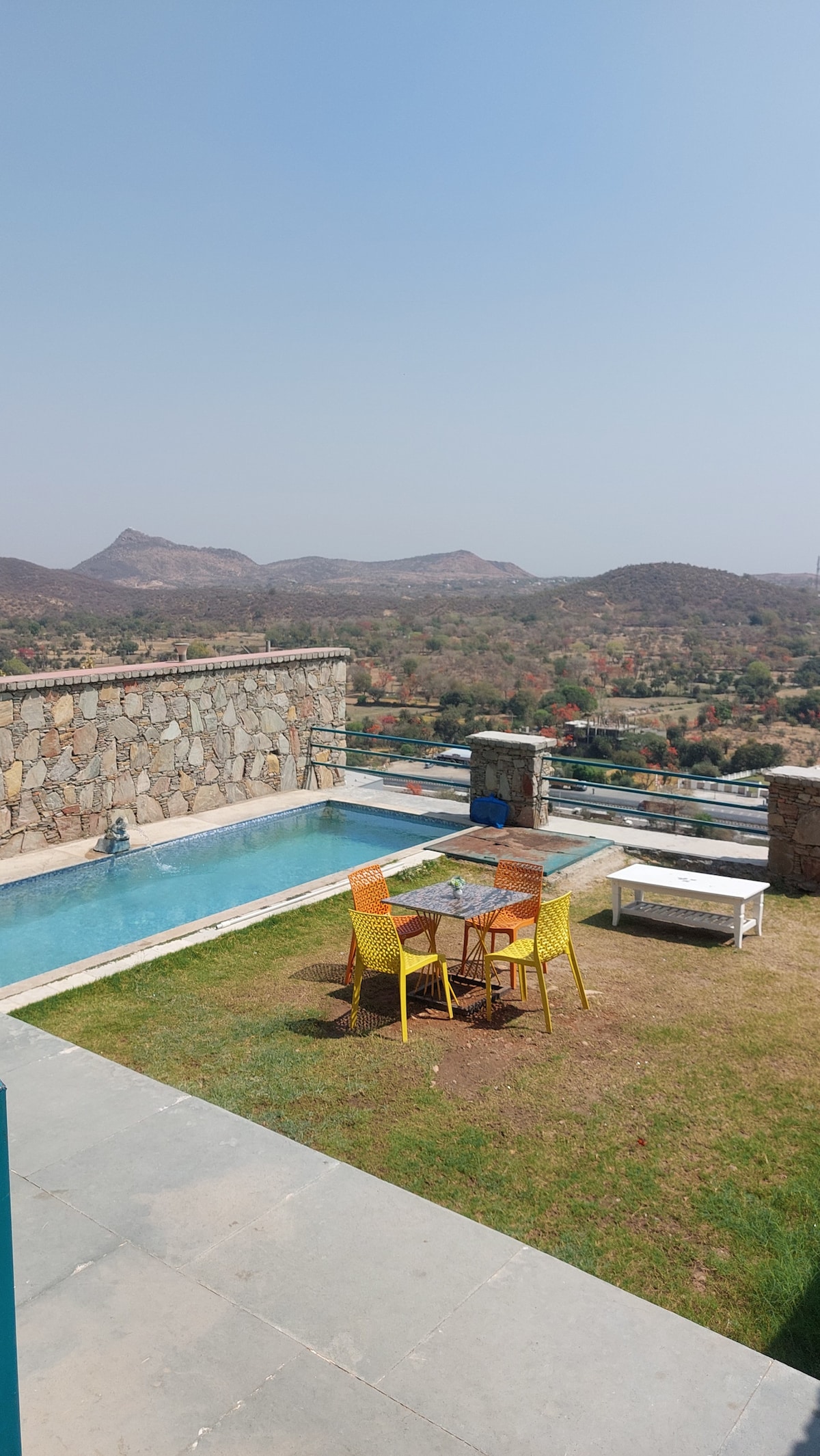 An outdoor seating area is showcased near a private pool, surrounded by lush greenery and hillside views. A glass-top table sits adjacent to colorful yellow chairs on a grassy patch, complemented by a white bench. The stone wall offers a rustic backdrop to the serene setting.