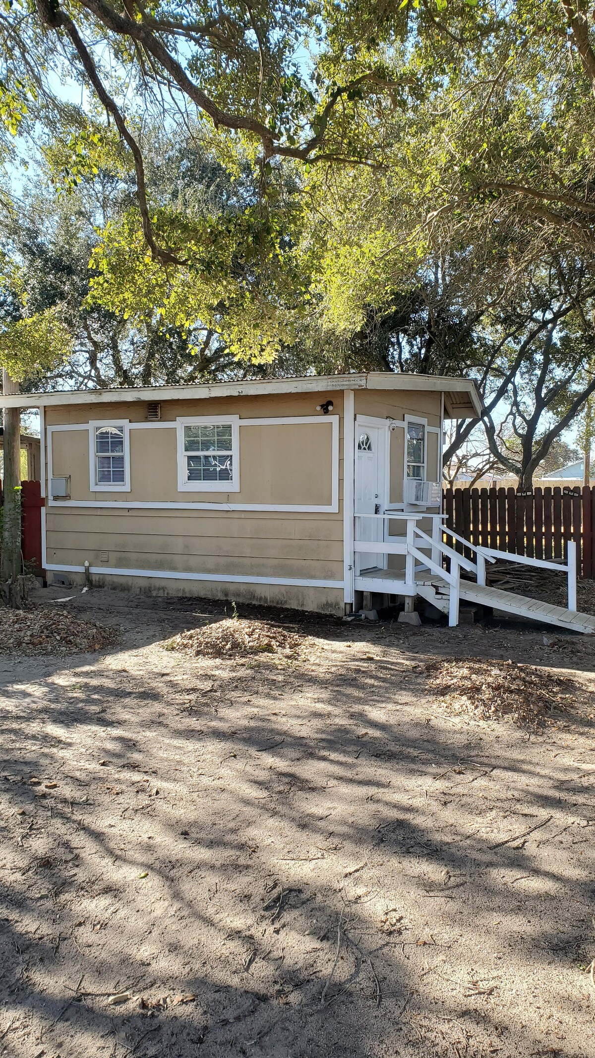 A small tan cottage with a white stairway leading to the front door is set amidst a spacious yard. Sunlight filters through the trees, casting gentle shadows on the ground. The property is enclosed by a wooden fence, enhancing privacy.