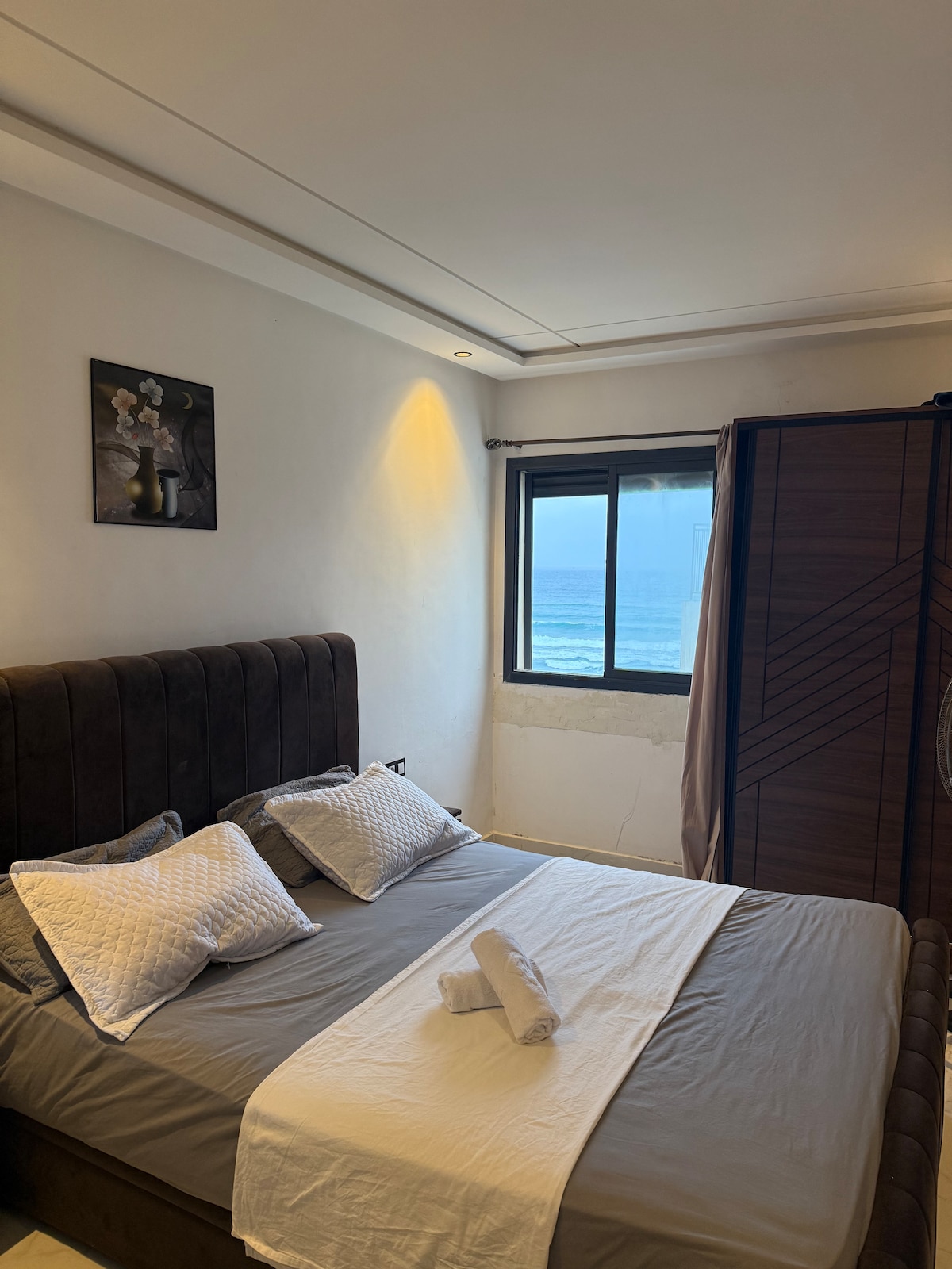 A sleeping area is equipped with a spacious bed featuring neutral-colored linens and decorative pillows. A window provides a view of the ocean, while natural light brightens the room. A modern wardrobe is positioned nearby, adding functionality to the aesthetic.