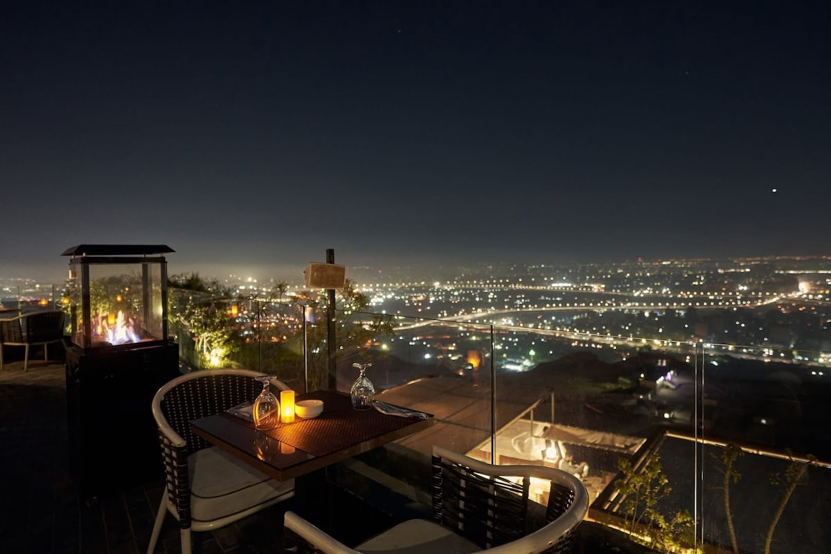A rooftop dining area is showcased with cozy seating arrangements. A table is set for two, featuring a soft glow from a candle. A glass barrier provides unobstructed views of the city skyline, illuminated by countless lights against the night sky.
