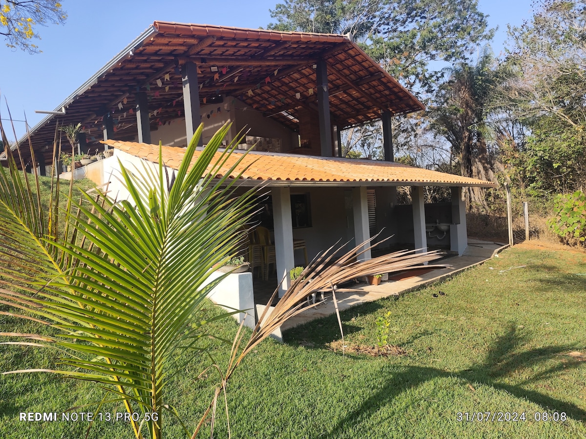 The exterior view of the home showcases a single-story structure with a sloped roof. A spacious patio is visible, surrounded by well-maintained grass and tropical plants. Bright sunlight illuminates the area, highlighting the garden's natural beauty and inviting ambiance.