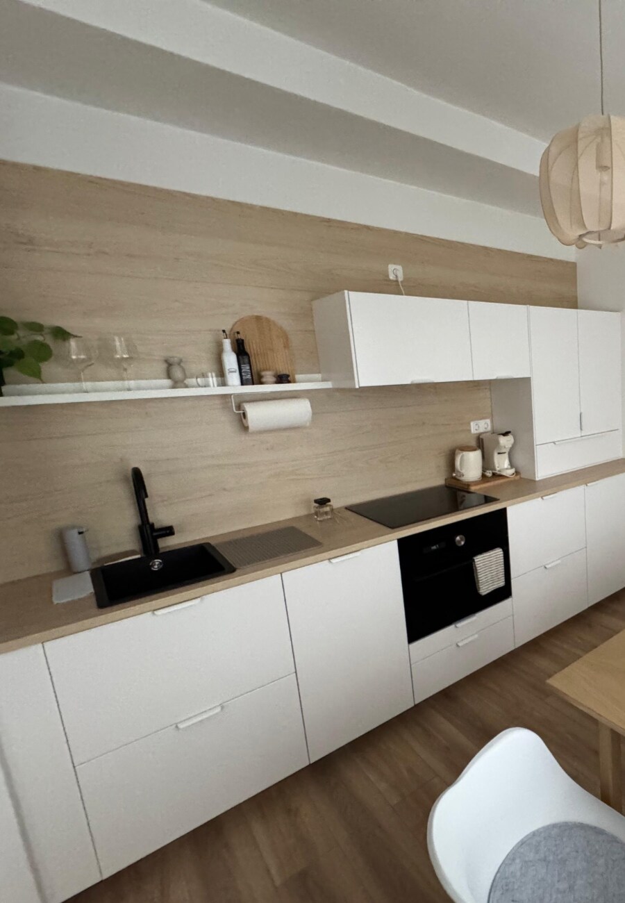 The kitchen area features modern white cabinetry against a light wood wall. Equipped with an efficient black sink and a built-in oven, the countertop includes space for meal preparation. Simple decor elements offer a clean and functional atmosphere, encouraging a comfortable cooking experience.