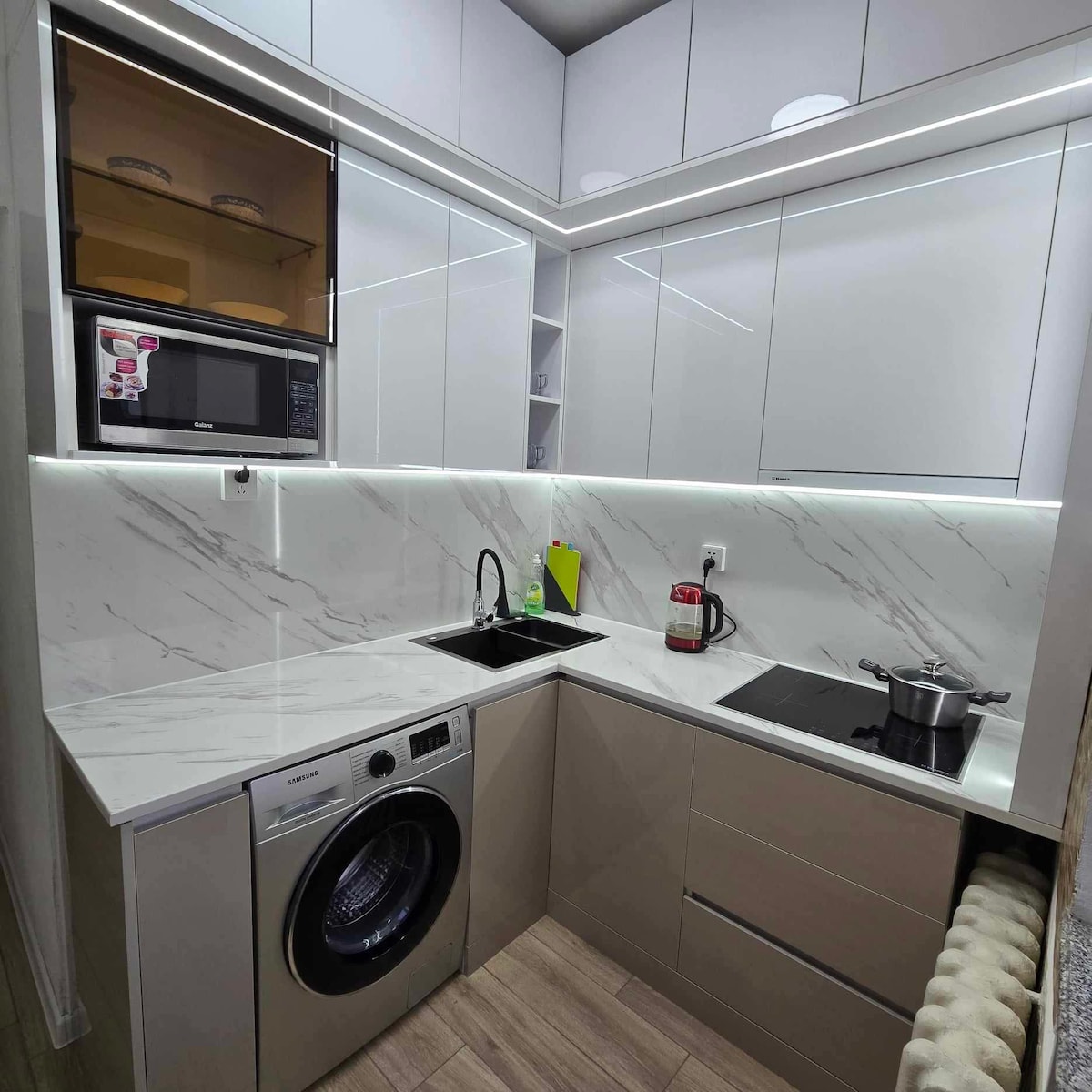 A modern kitchen features sleek cabinets with a glossy finish and a marble countertop. Appliances include a washing machine, microwave, and a cooking surface. Under-cabinet lighting illuminates the space, enhancing the functionality while a kettle and a pot are visible on the counter.