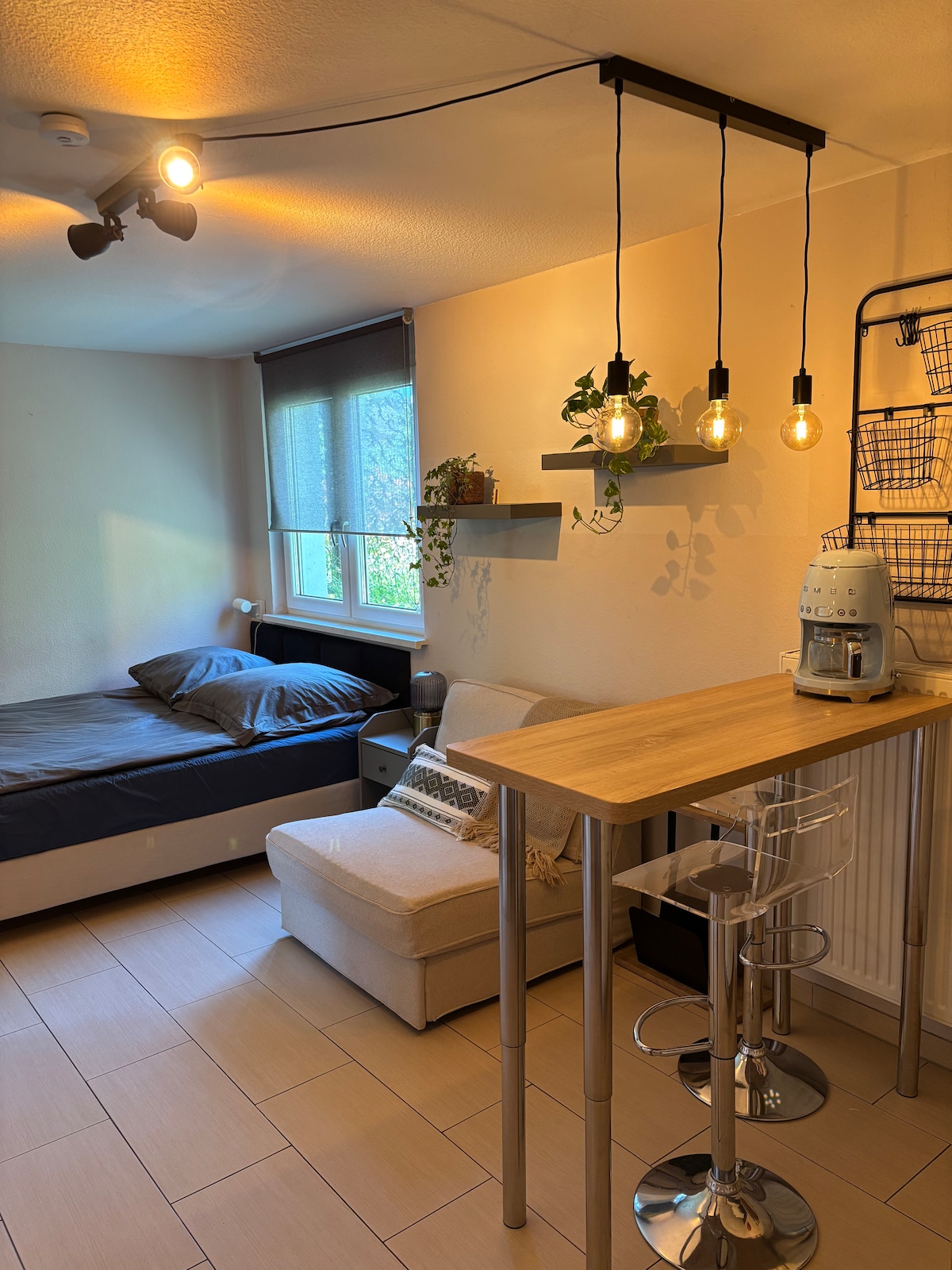 A cozy apartment corner features a spacious bed with a simple headboard, positioned against the wall. A soft couch sits nearby, complemented by a small dining table with tall stools. Three pendant lights hang above, illuminating the space, while a window reveals natural light.