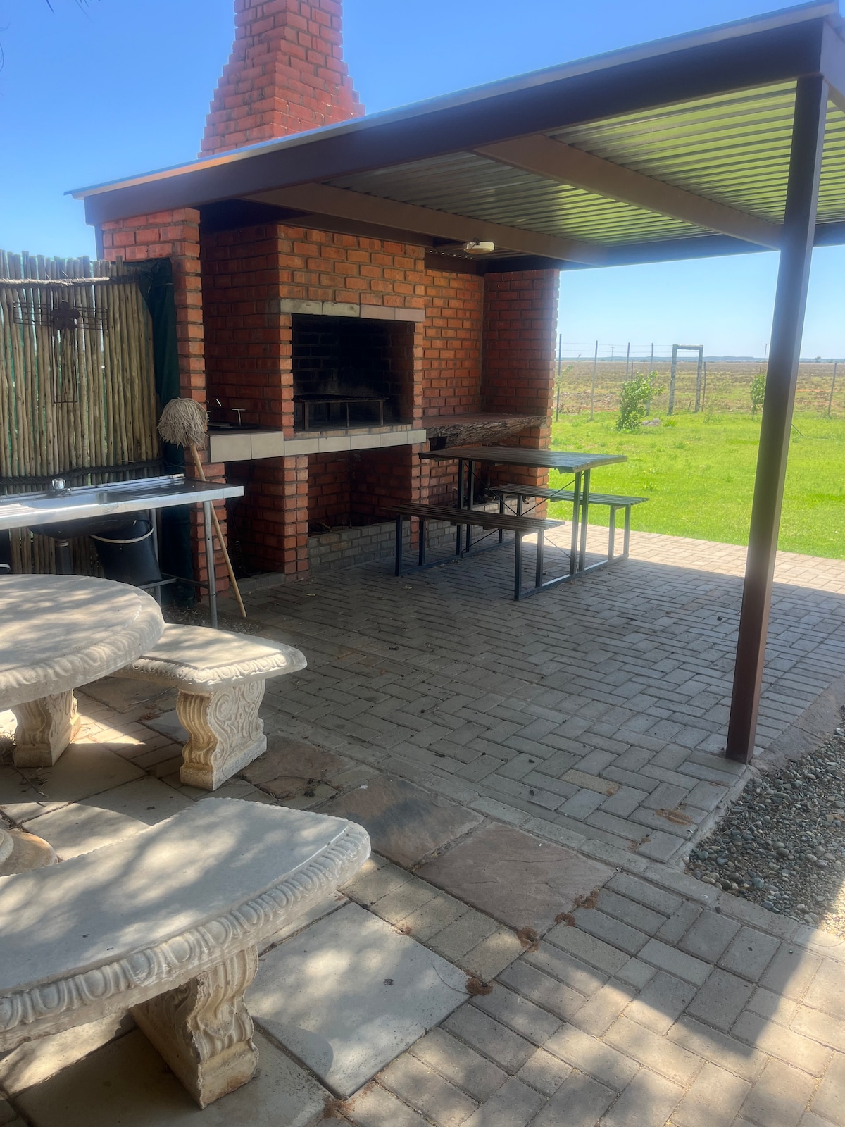 An outdoor cooking area features a built-in brick barbecue and a covered dining space. A light stone table with matching benches is visible, surrounded by paved flooring. Lush green grass is seen in the background, adding to the serene atmosphere of the farm setting.
