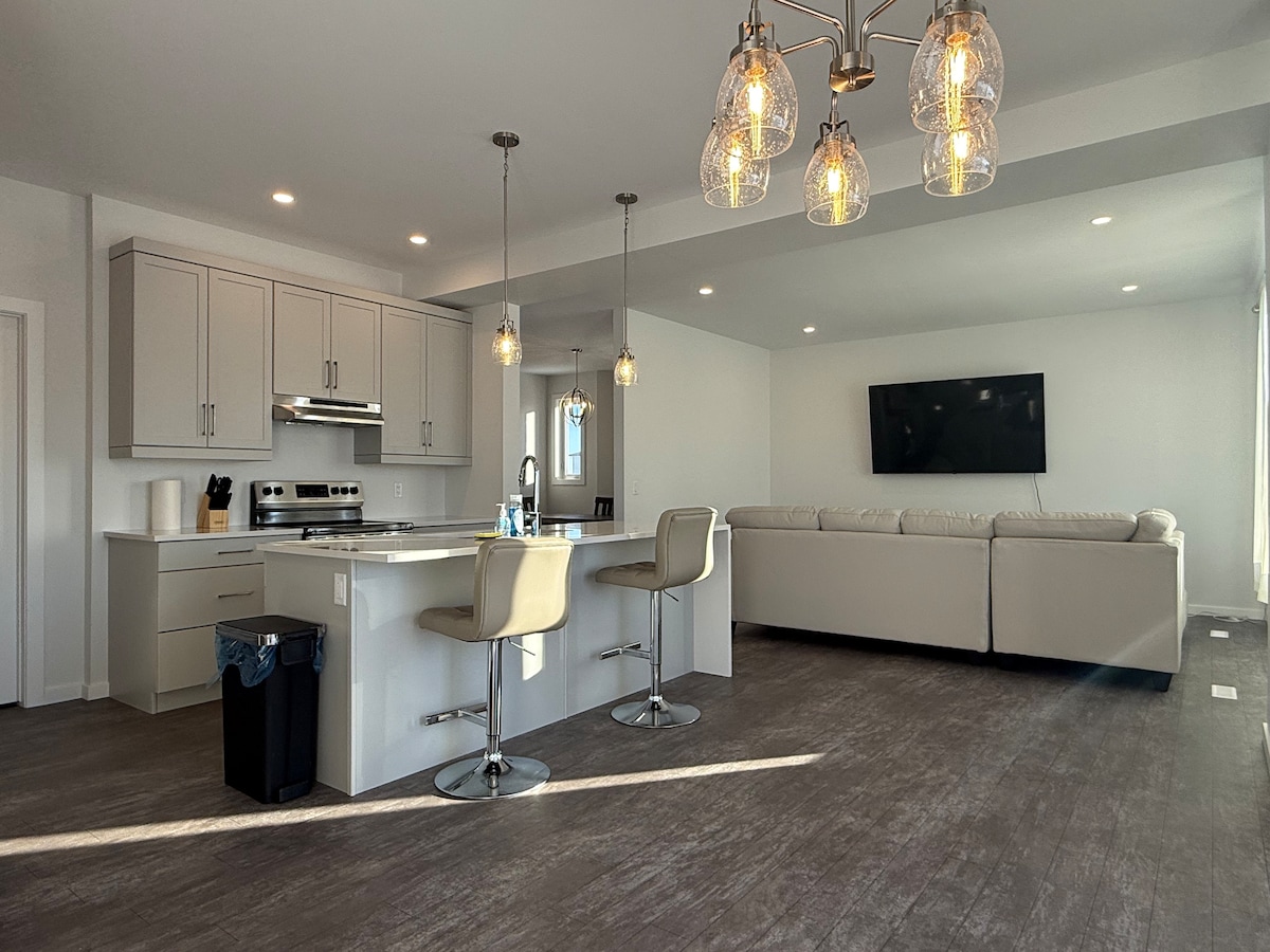 An open-concept kitchen and living area features modern cabinetry, stainless steel appliances, and a large island with seating. Natural light fills the space through oversized windows, with a comfortable sectional sofa and a wall-mounted smart TV creating a welcoming environment for relaxation.