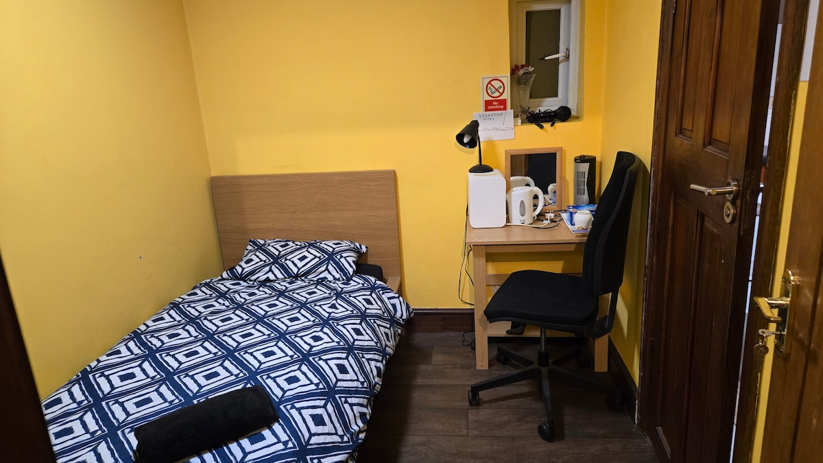 A small room is shown with bright yellow walls. A narrow bed covered with a geometric-patterned comforter is positioned against one wall. A compact desk with a white kettle and cups sits nearby, along with a black chair. A mirror is mounted above the desk.