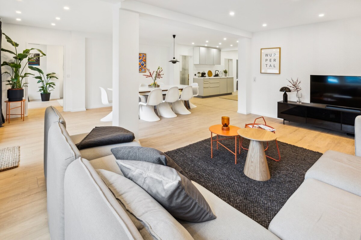 A spacious living area features a light-colored sectional sofa and a round coffee table atop a dark area rug. The open layout connects to a dining area with sculptural chairs, while a modern kitchen is visible in the background. Large windows provide ample natural light.