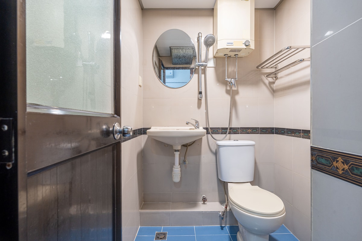 The bathroom features a compact design with a toilet and a sink positioned against light-colored tiled walls. A circular mirror is mounted above the sink, and a shower area is visible, separated by a glass door. Blue tiles offer a contrasting accent.