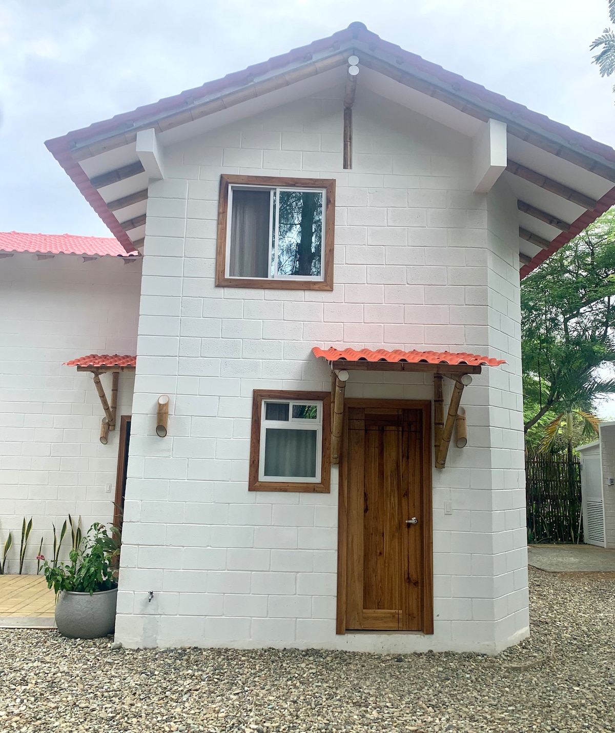 A two-story beachfront loft is displayed, featuring a light-colored exterior with a wooden door and window frames. The charming roof has red tiles, and small accent windows complement the design. Surrounding greenery enhances the peaceful environment, creating an inviting entrance.