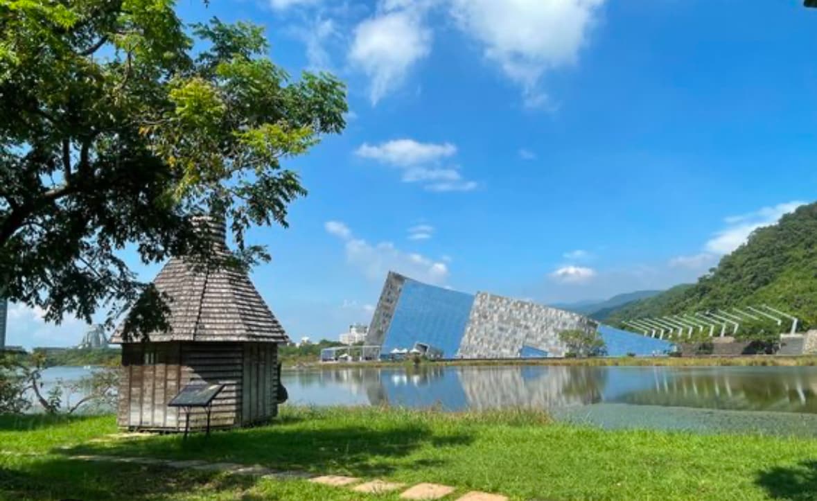 A serene landscape features a reflective body of water bordered by green foliage. A small wooden structure is positioned in the foreground, while a modern angled building emerges from the water's edge, surrounded by rolling hills and a clear blue sky.