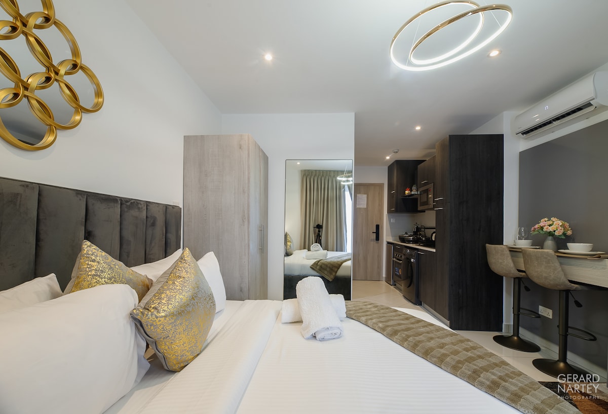 A cozy studio apartment features a neatly made bed with golden accent pillows and a soft blanket. Modern cabinetry is visible in the background, along with a compact cooking area. A dining table for two is positioned near the kitchenette, complemented by soft lighting from a circular fixture.