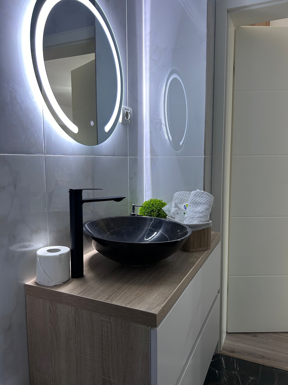 A modern bathroom features a sleek dark bowl sink atop a wooden vanity. A circular mirror illuminated by soft light is mounted above. A roll of tissue and a small plant add subtle touches to the clean, minimalist design.