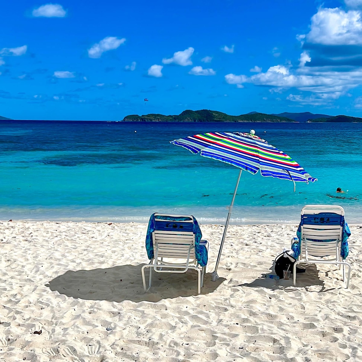 Two beach chairs with colorful striped umbrellas are positioned on a sandy shore. The clear turquoise waters stretch out in front, with gentle waves lapping at the beach. Islands are faintly visible in the background under a bright blue sky dotted with clouds.