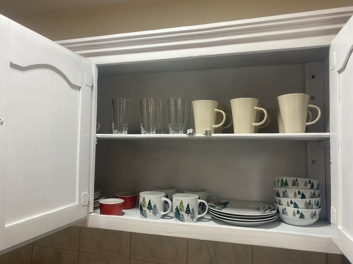 A white cabinet is opened to reveal neatly arranged dishware. The upper shelf holds various glasses and cream-colored mugs, while the lower shelf features a variety of plates and mugs adorned with winter-themed designs, creating an organized dining space.