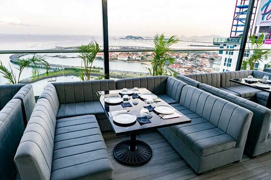 A modern dining area features sleek gray sofas arranged around a black table, designed for comfort and conversation. Panoramic views of the city and bay are visible through large glass windows, highlighted by the soft glow of the evening sky.