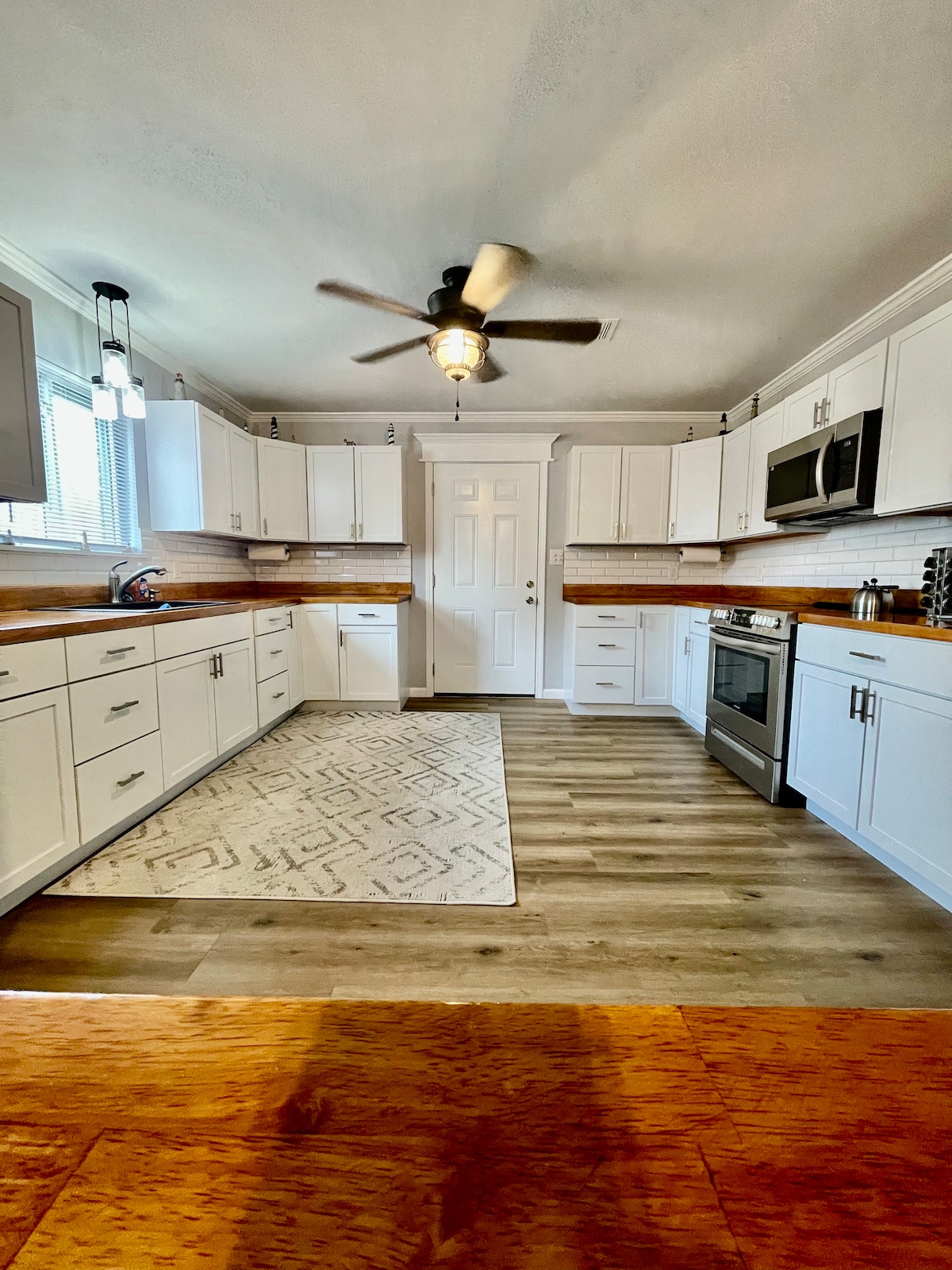 A bright kitchen features white cabinetry with ample storage, complemented by wood accents. Stainless steel appliances, including a microwave and oven, are present. A patterned area rug is positioned on the floor, and natural light enters through a nearby window, enhancing the space's inviting ambiance.