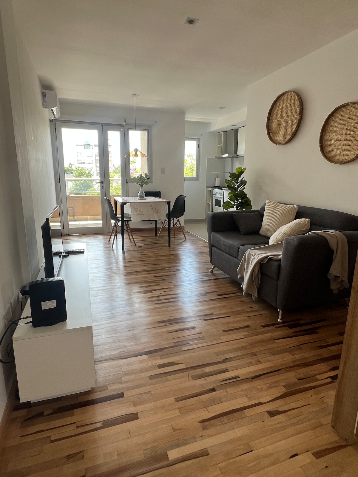 An open living area features a gray sofa with light-colored cushions, positioned beside a white media console. A dining table is set for two, with a view of outdoors visible through large glass doors. Wooden flooring adds warmth to the space.