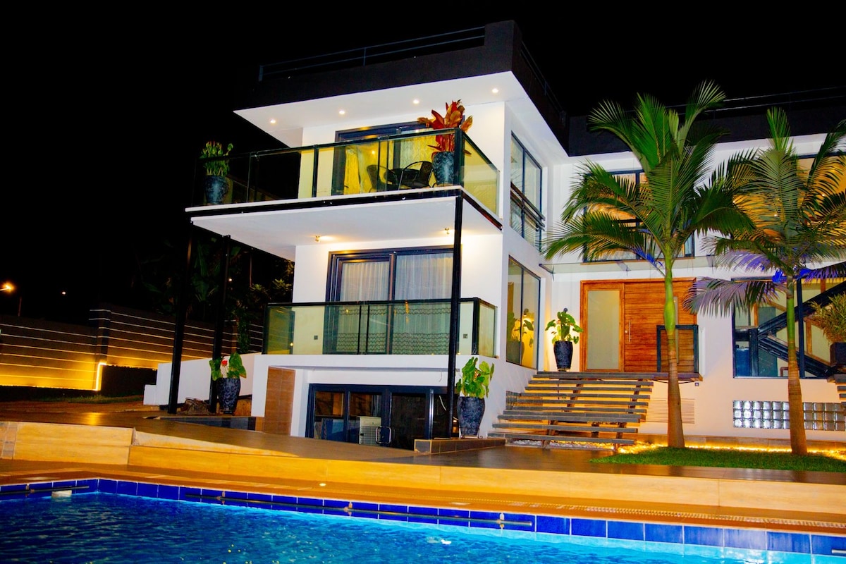 A modern two-story house is depicted at night, featuring large glass balconies and vibrant landscaping. Soft lighting highlights the exterior, while a swimming pool with clear blue water is visible in the foreground, reflecting the house's architecture.