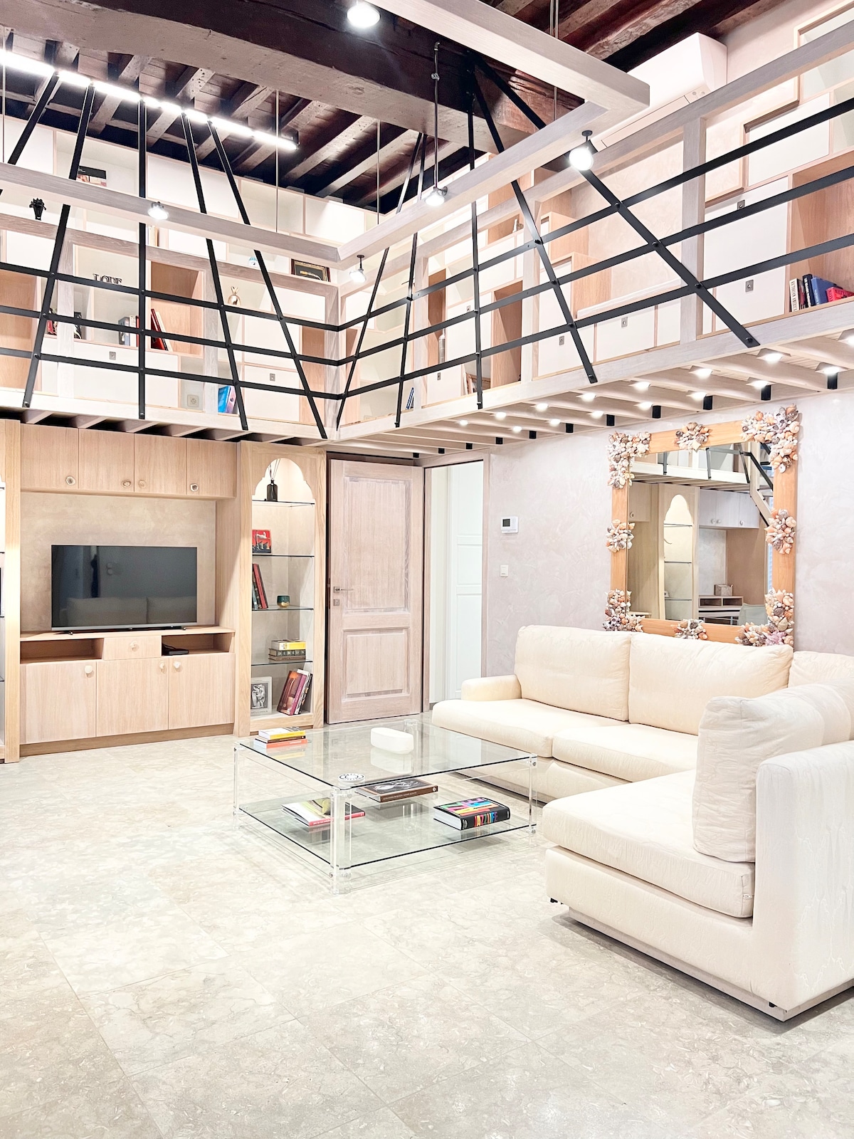 The living area displays a spacious and elegant design, featuring a light-colored sofa positioned in front of a modern entertainment unit. A glass coffee table rests on the marble flooring, while built-in shelves showcase books and decorative items. Ambient lighting highlights the pleasing architectural details.