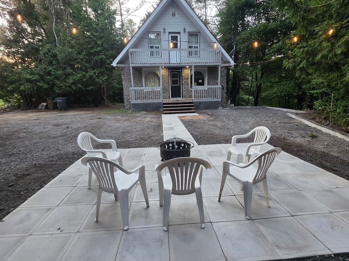 An outdoor patio area is featured, consisting of a light-colored paved surface with multiple white chairs arranged around a central fire pit. The charming rustic house is visible in the background, surrounded by greenery and illuminated by string lights.