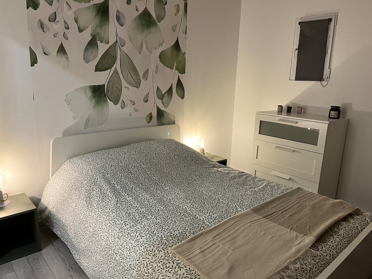 A spacious bed is positioned against a wall adorned with a leafy design. Soft lighting from two bedside lamps creates a gentle ambiance. A light-colored throw is neatly placed at the foot of the bed, complemented by a sleek dresser against the opposite wall.