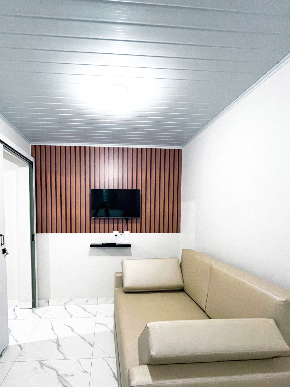 A small seating area is included, featuring a light-colored sofa positioned against a wooden accent wall. A flat-screen television is mounted on the wall opposite the sofa, while a small shelf is visible below the TV. Bright overhead lighting provides ample illumination.