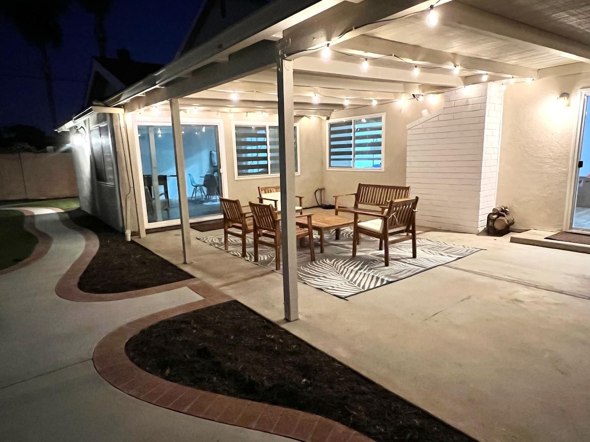 An outdoor patio is illuminated by string lights, creating a cozy ambiance in the evening. Wooden chairs and a table are arranged on a patterned rug, with a smooth concrete floor leading to the spacious yard and large windows that offer views into the home.