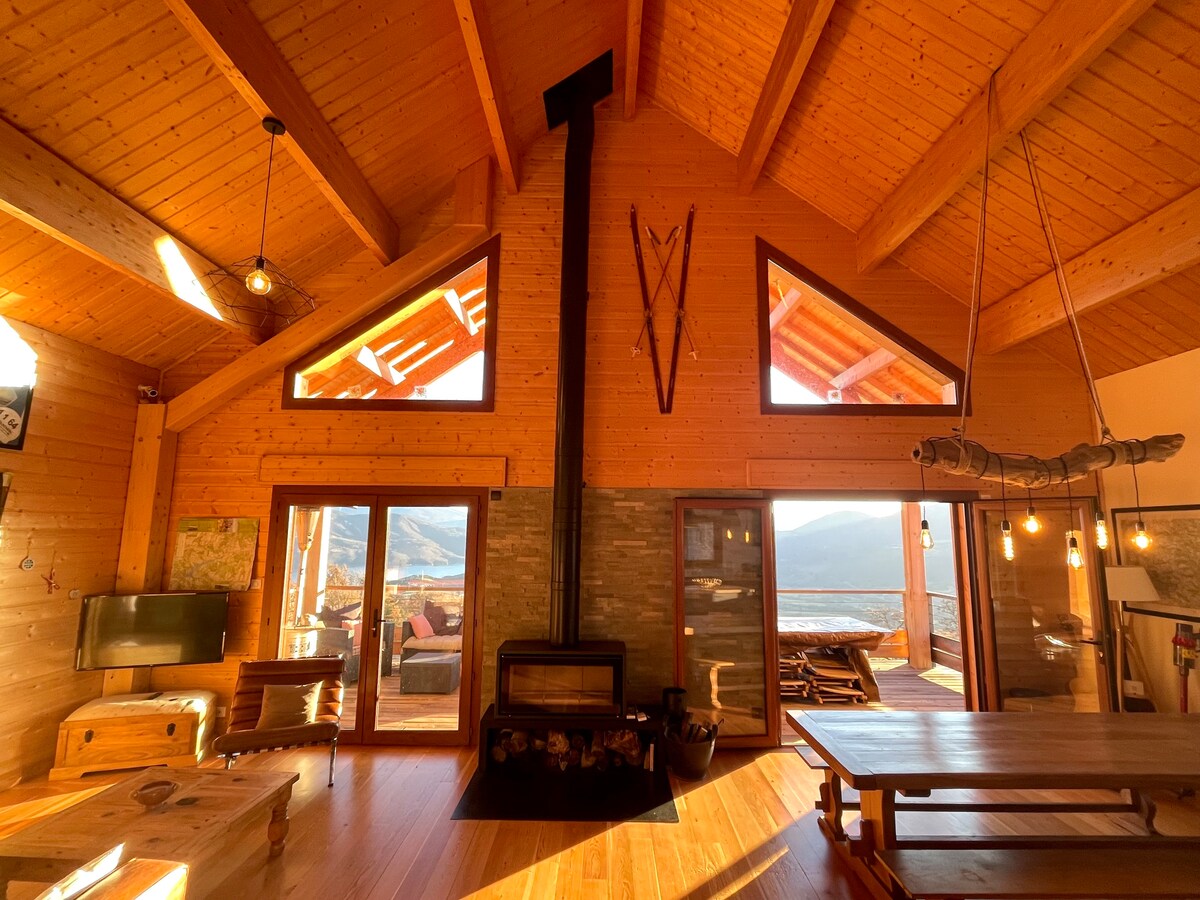 The spacious living area features high wooden ceilings adorned with beams and large windows that bring in natural light and provide expansive views. A central wood-burning stove is situated between two seating areas. A dining table is positioned to the right, enhancing the open layout.