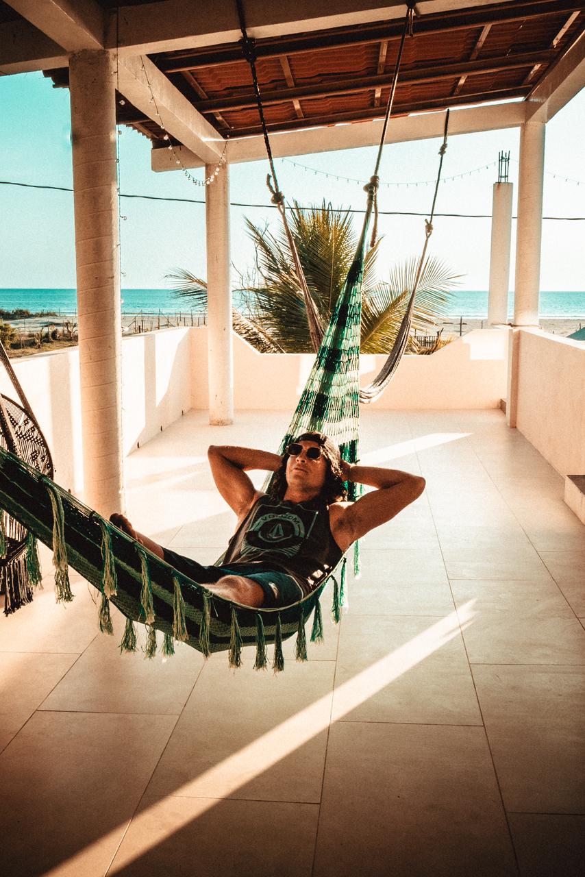 A shaded terrace features hanging hammocks, inviting relaxation with a view of the ocean. Soft sunlight filters through the open space, highlighting the tranquil atmosphere created by palm trees swaying in the gentle breeze.
