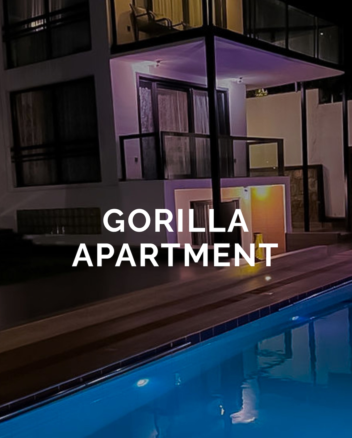 An elegantly designed apartment building is shown at night, illuminated by soft lighting. A serene swimming pool reflects the lights of the structure, creating a calming ambiance. The outdoor terrace is visible, which overlooks the pool area.
