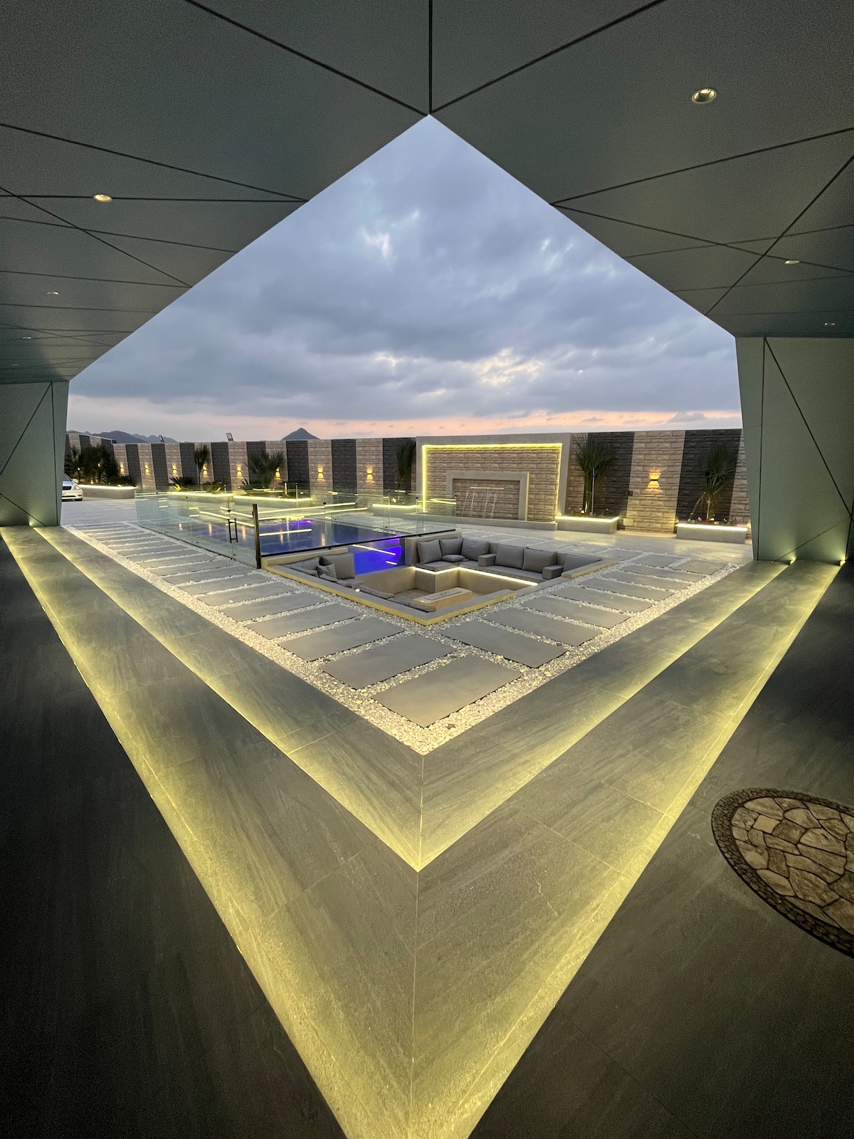 A spacious outdoor area is framed by modern architecture. Soft lighting outlines the geometric pathways leading to a landscaped space with seating and a pool. The sky displays varying shades of grey and blue, creating a calming backdrop for the luxurious environment.