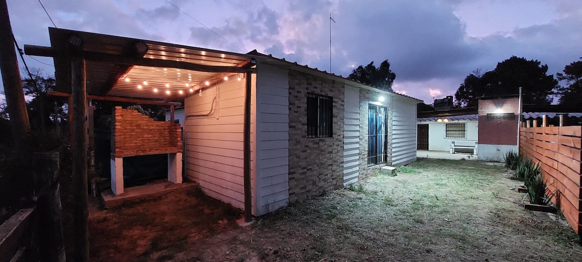 An exterior view of a newly constructed casita is shown, with a covered grill area illuminated by string lights. The building features a combination of brick and siding, with large windows providing a glimpse of the interior. A grassy area surrounds the structure, enhancing the outdoor space.