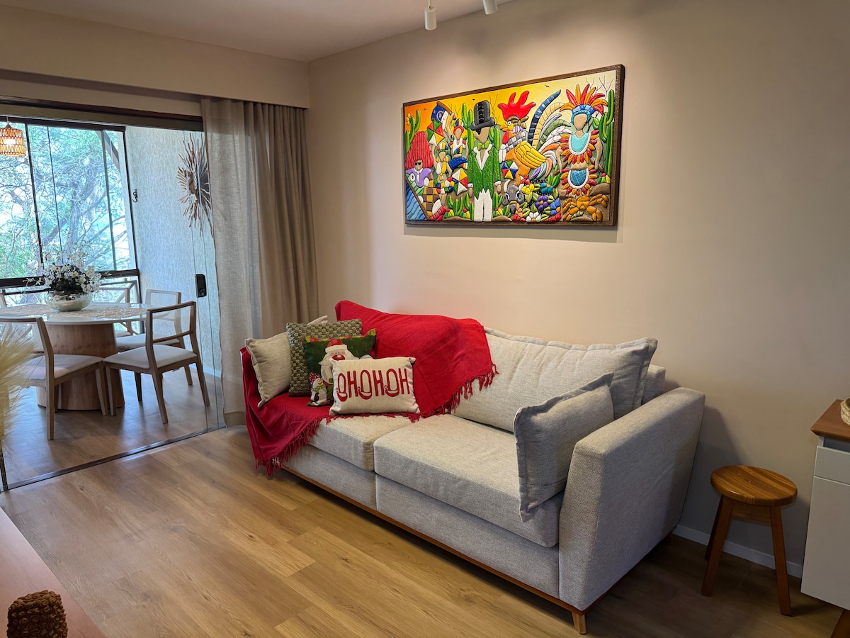 A light-filled living area features a comfortable gray sofa adorned with colorful cushions and a red throw blanket. A wooden stool complements the modern design, while a dining area with natural light is visible through large glass doors.