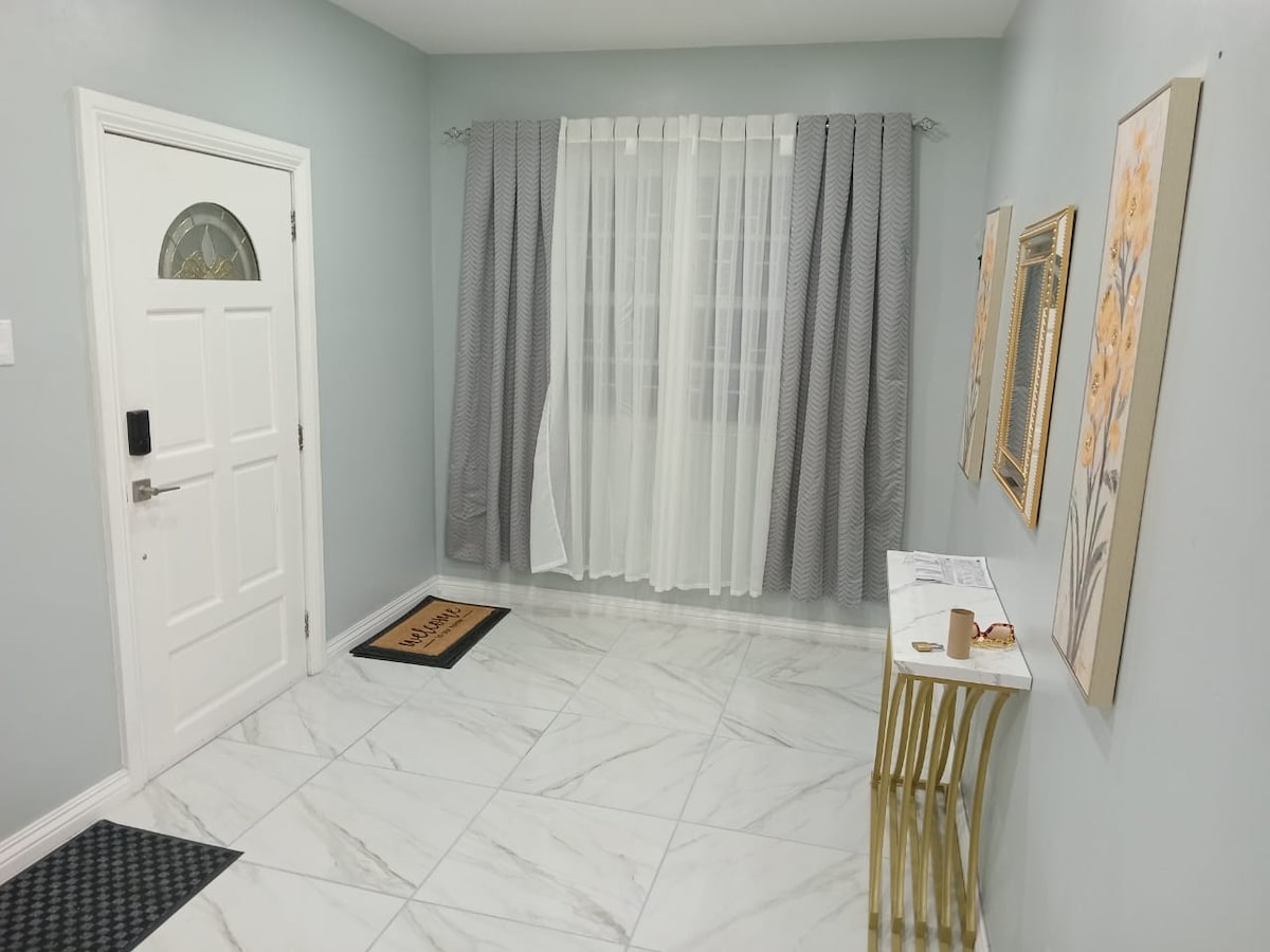 An entryway features elegant marble flooring and light gray walls. A white door stands at the entrance, flanked by sheer white curtains. Two decorative gold-framed mirrors are mounted on the wall, and a small console table adds a functional touch. A welcome mat lies at the door.