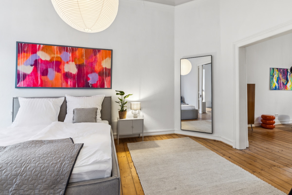 A well-lit bedroom features a spacious bed dressed in crisp white linens, with decorative pillows arranged neatly. A large mirror reflects the room, and a colorful wall art piece adds vibrancy above the bed. A plant and bedside lamp are positioned on a nightstand beside the bed.