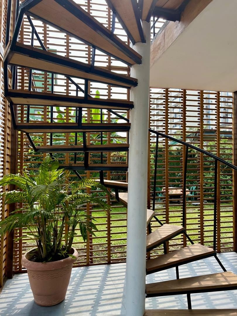A spiral staircase is framed by wooden slatted walls, allowing natural light to filter through. A potted plant is positioned at the base of the stairs, complementing the outdoor greenery visible beyond. The setting offers a balance of indoor and outdoor elements.