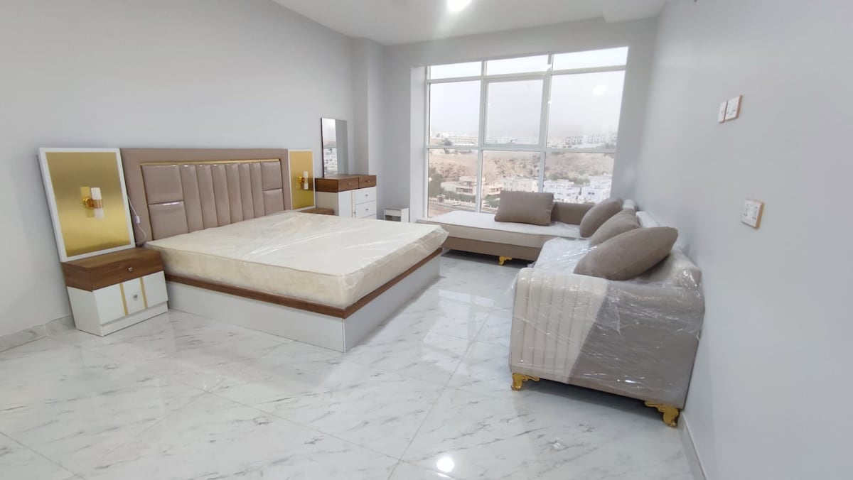 A spacious bedroom features a large bed with a stylish headboard and a minimalist design. A comfortable sofa sits next to a window, allowing natural light to fill the room. Nightstands with lamps and a modern dresser add functionality to the space.