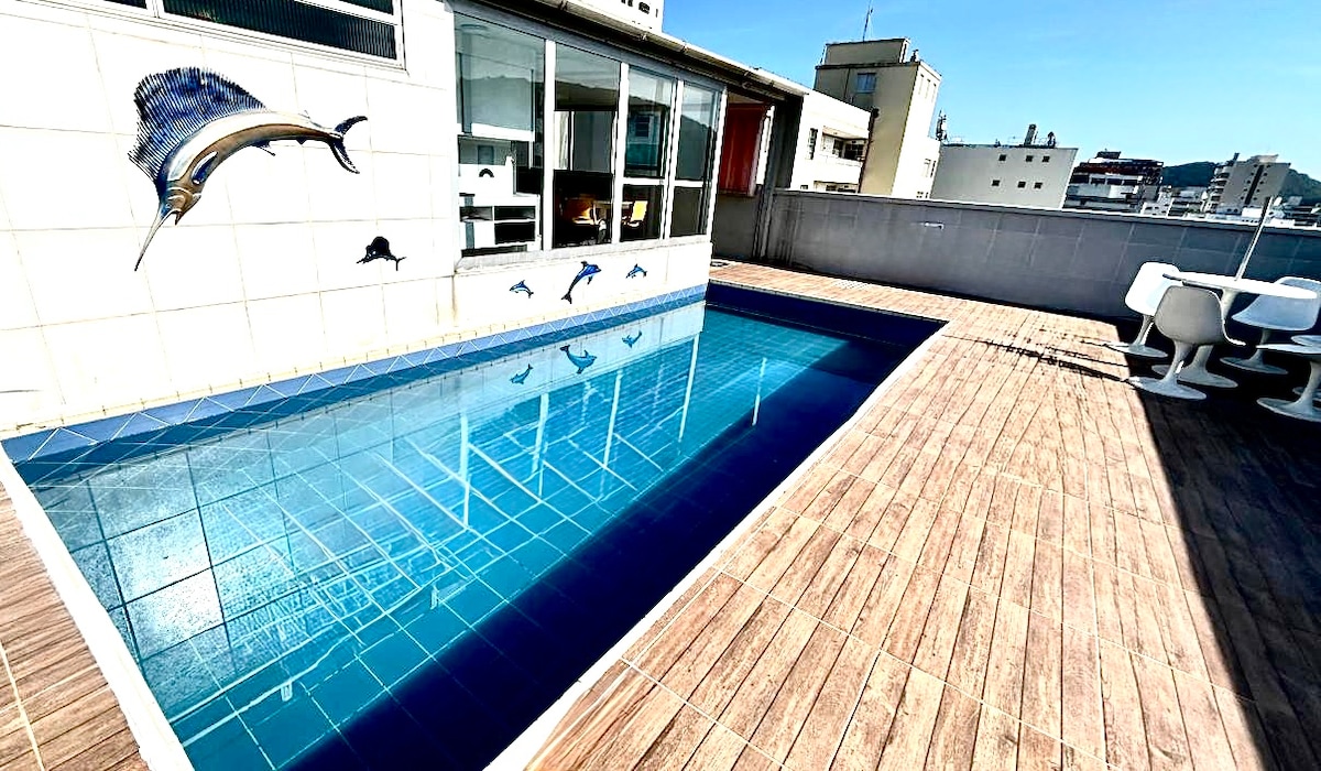 A private pool is set on a spacious wooden deck, featuring a serene blue tile design. The backdrop includes large windows that allow natural light to fill the area. The walls are adorned with marine artwork, contributing to a relaxed outdoor space.
