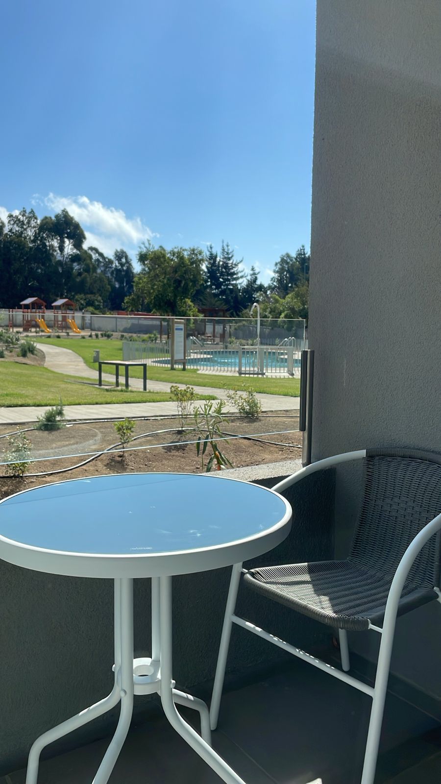 A small outdoor balcony features a round table and a single chair, overlooking a landscaped area. In the background, a swimming pool is visible along with recreational facilities set amidst greenery. Clear skies enhance the inviting atmosphere.