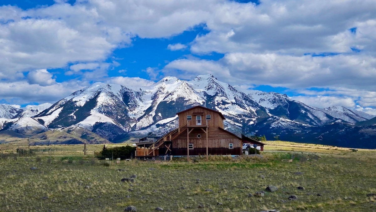 The two-story wooden house is situated against a backdrop of majestic snow-capped mountains under a partly cloudy sky. Expansive grassy fields stretch around the property, providing a sense of openness and connection to the natural landscape.
